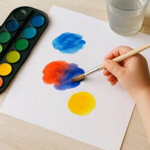 A child’s hand mixing red and blue watercolor paint on white paper, with a paintbrush, watercolor set, and water cup beside it.
