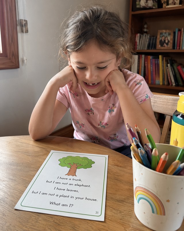 Candid snapshot of a child aged 6 with a gap-toothed smile sitting at a small round wooden desk or kitchen table, wearing a light pink t-shirt with a