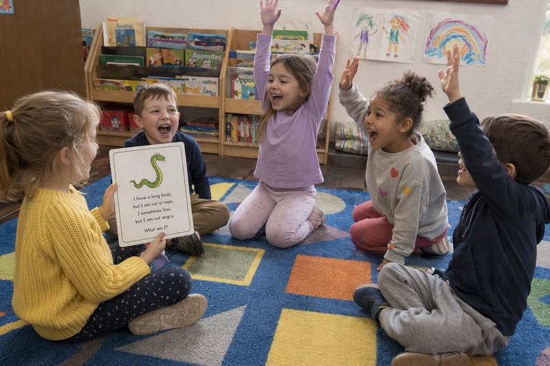 Candid snapshot of five children aged 5 to 6 sitting in a loose circle on a colourful patterned classroom rug with geometric shapes on it, one child i