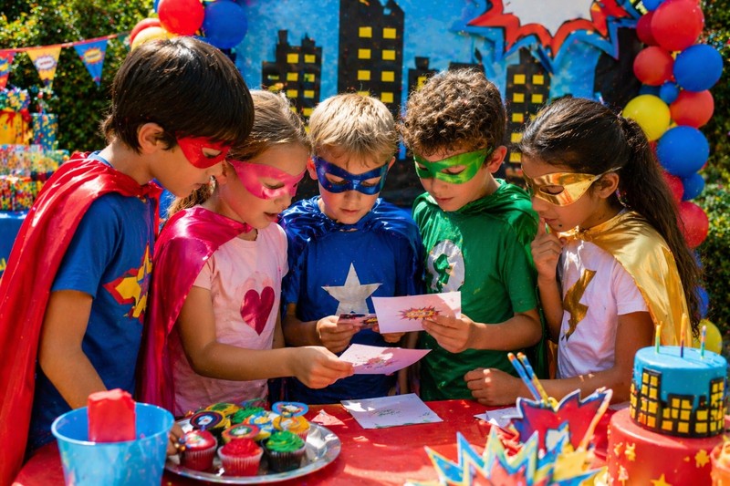 Children wearing superhero capes and masks solving riddle cards together at a colourful superhero birthday party