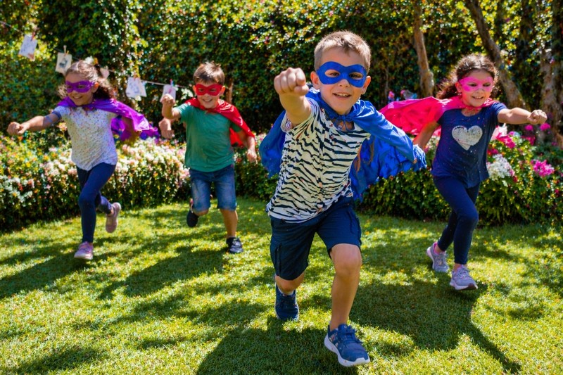 Children running through a garden wearing homemade superhero capes, pretending to fly while searching for riddle clue cards
