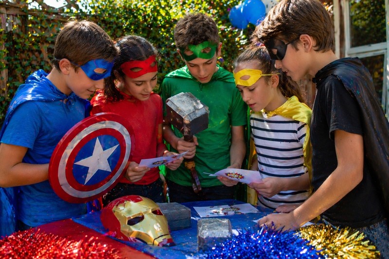 Older children examining superhero gadget props like a cardboard shield and toy hammer while solving riddle cards at a party table