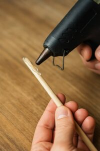 Nahaufnahme: ae Hand trägt with aer Heißklebepistole Kleber auf aen Holzstab auf – the erste Schritt bein the crafts aes magic wands for Kinthe with DIY-Materialien