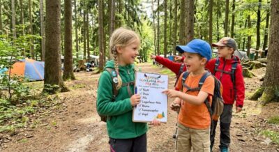 Three children sitting on a picnic blanket at a forest campsite holding white printable scavenger hunt sheets during a fun camping scavenger hunt for kids.