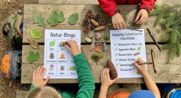 Top-down view of children playing nature bingo and checking off a camping scavenger hunt list on a wooden picnic table outdoors, featuring found forest items like pinecones and leaves.