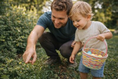 Parent kneeling at child level pointing at something in the grass while playing an outdoor scavenger hunt with a toddler holding a basket, natural light, realistic DSLR-style photo