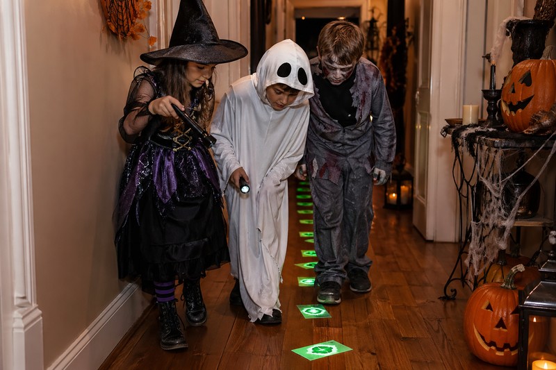 Outdoor Halloween hunt at dusk: children with flashlights exploring a decorated backyard, illuminated pumpkins on path, cobweb fences, clue note taped