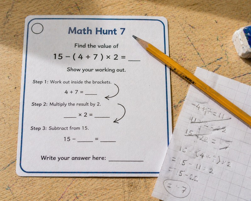 Close-up snapshot of a printed math scavenger hunt clue card lying flat on a light natural wood school desk, the card showing a simple multi-step equa