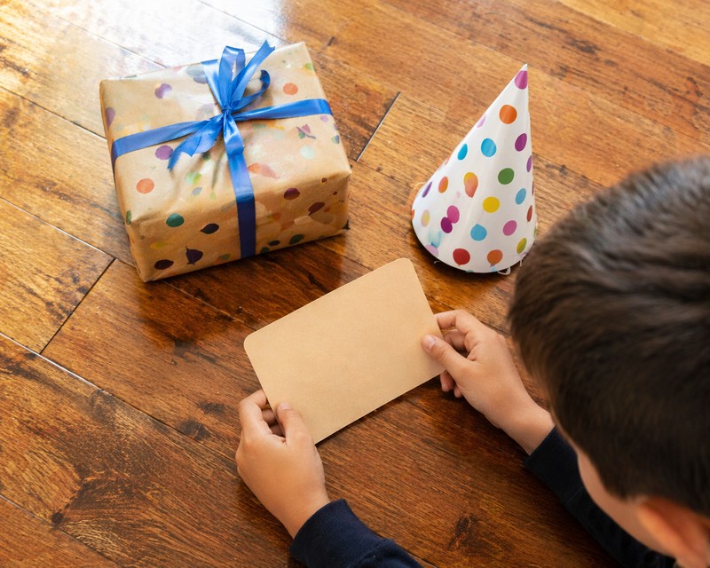 Overhead snapshot of a birthday scavenger hunt clue card on a wood floor next to a wrapped birthday present and a party hat, a child’s hands picking u