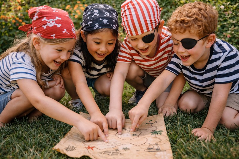 Outdoor pirate treasure hunt: three children in pirate gear exploring a backyard — behind garden shed (ship mast), under a large potted plant (palm tr