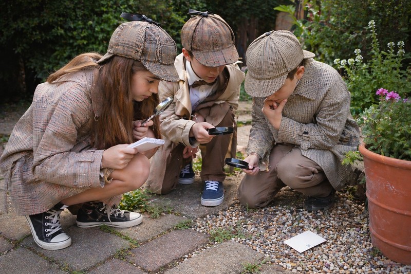 Candid snapshot of three children aged 7 to 10 in detective costumes — magnifying glasses, deerstalker hats, one with a notepad — crouching on a garde