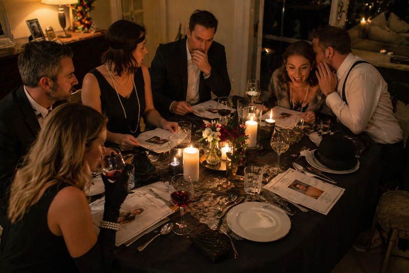 Candid snapshot of six adults at a dinner table set for a murder mystery evening, printed character role cards at each place setting, one guest leanin