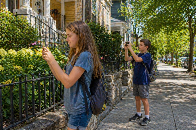 Neighborhood photo hunt: two older children navigating a suburban street with smartphones, taking photos of architectural details and nature finds, co