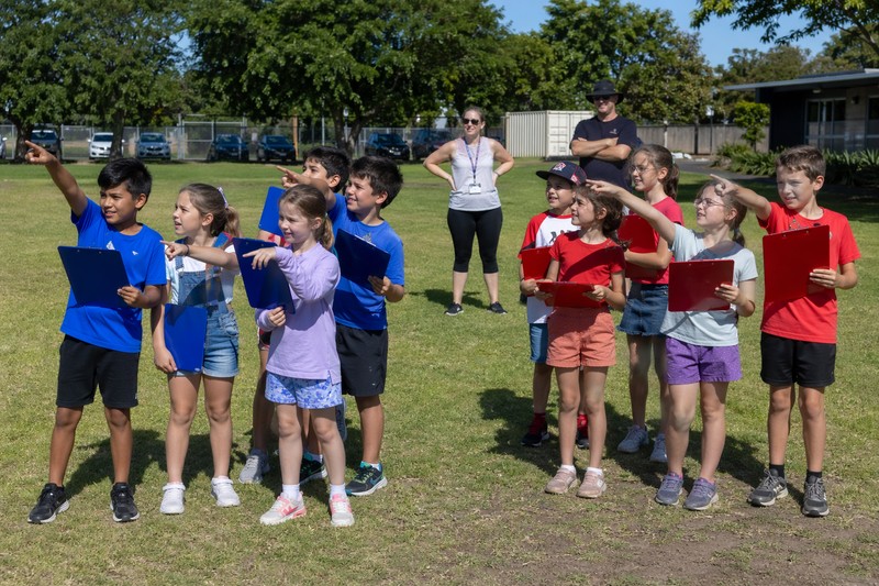 Candid snapshot of twelve children aged 6 to 11 split into two teams on a school playing field, each team holding matching colored clipboards and poin